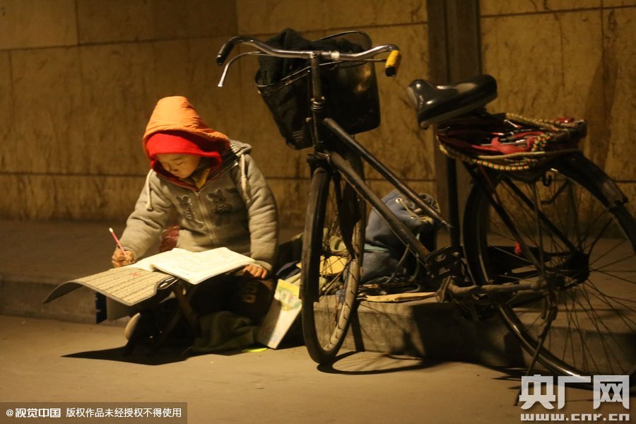 鄭州中原路京廣鐵路大橋下涵洞的馬路牙子邊，一位戴著紅色辣椒帽的小男孩趴在一個(gè)小馬扎上，上面鋪著一張車(chē)墊，在微弱的燈光下寫(xiě)作業(yè)